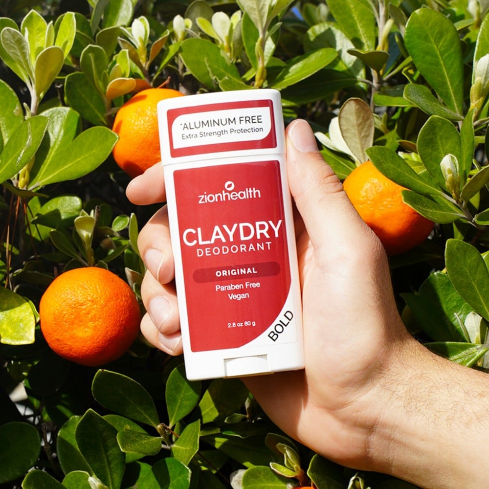 Hand holding CLAYDRY deodorant among orange trees.